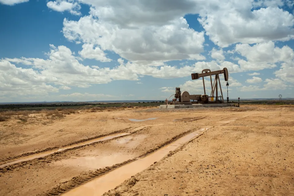 Distant view of brown oil pump jack in a flat desert with cloudy blue sky.